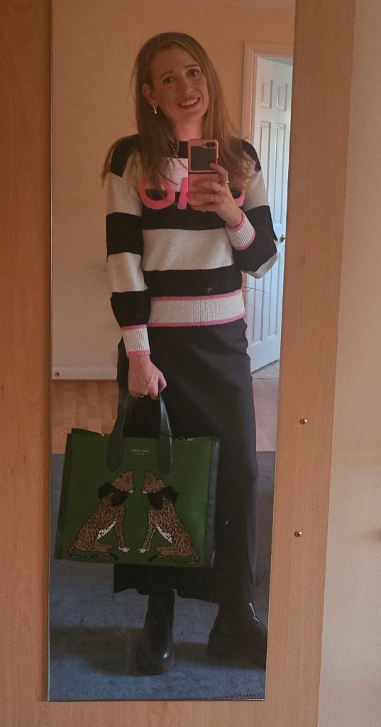 Woman taking a selfie in a mirror, wearing a striped sweater with 'CIIAO' text, a long black skirt, chunky black boots, and holding a green tote bag featuring two cheetahs..