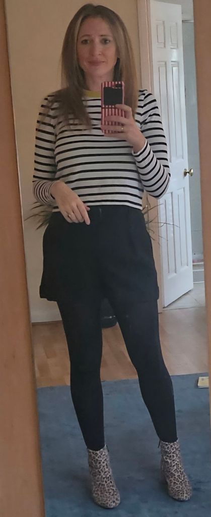 A woman in a striped long-sleeve shirt, black shorts and leopard print ankle boots posing in front of a mirror.