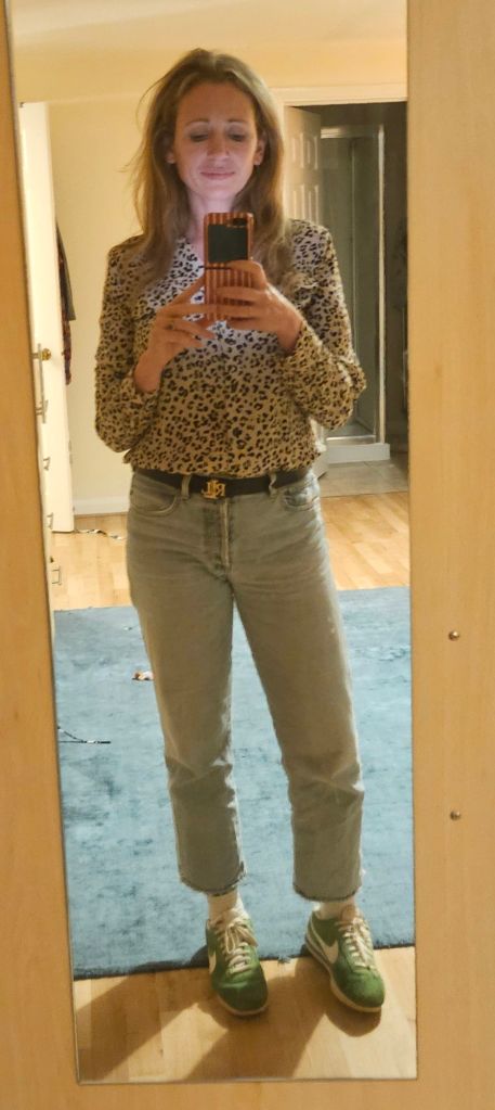 A person taking a selfie in a mirror, wearing a leopard print blouse, light blue jeans, and green sneakers, with a wooden floor and carpet visible in the background.